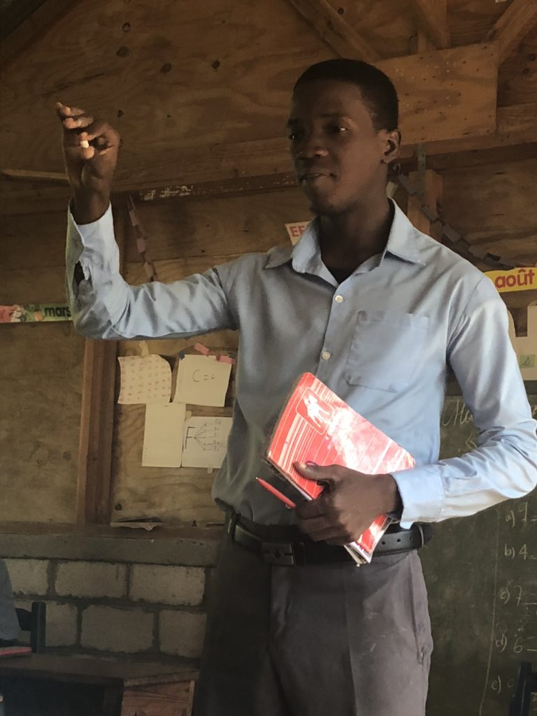 Arguens stands in a school classroom holding a book and gesturing with chalk to his students.